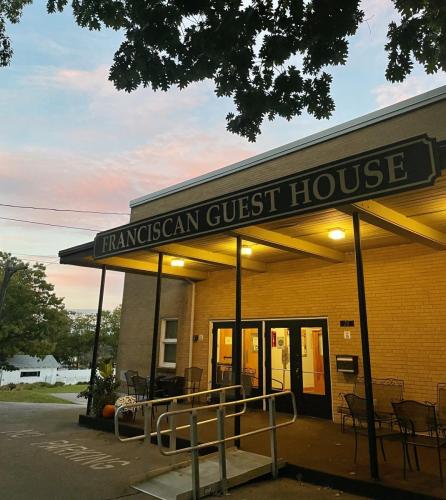 A tan building with an awning reading Franciscan Guest House lit by yellow lights from underneath.